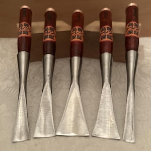 May include: Five wood carving chisels with dark brown wooden handles and metallic ferrules. Each handle features a circular orange label with black Japanese text. The steel blades vary in width and shape, ranging from narrow to wide flat edges.