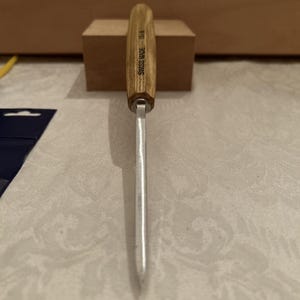 May include: A wood carving tool with a light brown wooden handle and a silver metal blade. The handle features the text SWISS MADE 1S/8 printed in black. The tool is positioned vertically, showing the narrow profile of the metal blade.