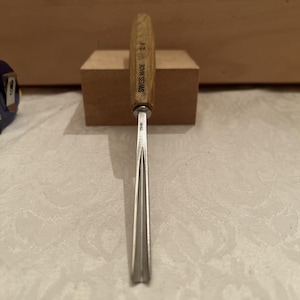 May include: A metal wood carving gouge with a light-coloured wooden handle. The handle is stamped with the text SWISS MADE and 12/1. The metal shaft features a curved blade profile and is stamped with the text 12/1 near the handle ferrule.