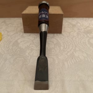 May include: A Japanese wood chisel with a dark brown wooden handle and a metallic ferrule. The handle features a blue circular sticker with white Japanese text that reads ローレンツ and 修復済. The tool has a long, black, tapered steel shaft.