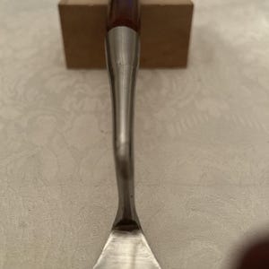May include: A metal chisel with a wooden handle. The handle is dark brown with a purple top. The chisel has a silver blade and a long, slender silver shaft. The tool is designed for woodworking and carving.