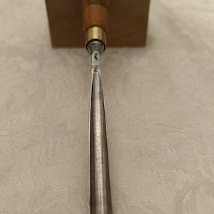 May include: A woodworking chisel with a wooden handle and a metal blade. The handle is a light brown color with a brass ferrule. The blade is a silver color and tapers to a point. The chisel is resting on a wooden block.