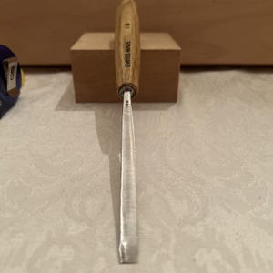 May include: A wood carving chisel with a light brown wooden handle and a silver metal blade. The handle is stamped with the text SWISS MADE and 1/8. The metal blade features a small engraved arrow symbol near the base of the handle.