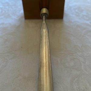 May include: A woodworking chisel with a wooden handle and a metal blade. The handle is a dark brown color, and the blade is silver. The chisel is positioned vertically against a wooden block.