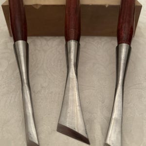 May include: A set of three woodworking chisels with dark reddish-brown wooden handles and metallic ferrules. Each tool features a long, slender steel blade with a sharp, angled cutting edge. The chisels are arranged vertically in a row.