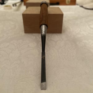 May include: A woodworking chisel featuring a long, dark brown wooden handle with a metal ferrule at the top. The metal shaft transitions from a cylindrical shape to a flat, narrow, dark-colored blade with a sharp, silver-toned cutting edge at the tip.