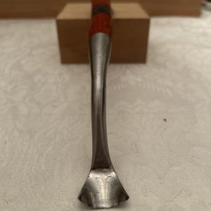 May include: A metal tool with a curved, scoop-shaped end and a long, slender neck. The handle features a section of reddish-brown wood with a dark, circular inlay. The tool is positioned vertically, showing its metallic sheen and smooth, curved design.