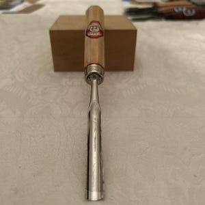 Puede incluir: Un formón de madera con un mango de madera de color claro y una hoja plateada. El mango tiene una etiqueta roja y blanca. El formón está colocado delante de una pequeña caja rectangular marrón. La herramienta está diseñada para carpintería.