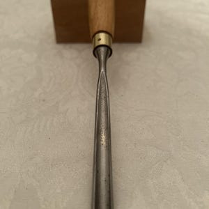 May include: A wood chisel featuring a wooden handle and a metal blade. The handle is light brown with the brand name "GREENLEE" printed on it. The metal blade is silver and tapers to a point.