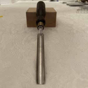 May include: A woodworking chisel with a dark wooden handle and a silver blade. The chisel is positioned vertically, with the blade facing down. The brand name "SWEARBY" is visible on the blade. The chisel is resting on a small, light brown wooden block.