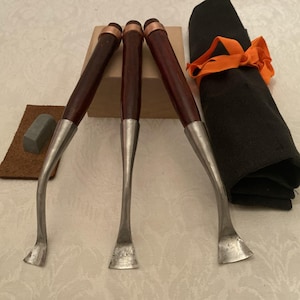 May include: Set of three woodworking gouges with dark wood handles, copper ferrules, and curved, silver-colored blades. A small sharpening stone and leather strop are visible, along with a black cloth tied with an orange ribbon.