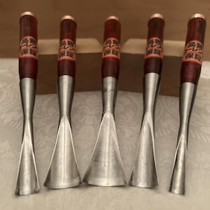 May include: Five wood carving gouges with polished metal blades and dark brown wooden handles. Each handle features a small orange label with black Japanese text. The tools are arranged vertically, showcasing varying blade widths and curvatures.