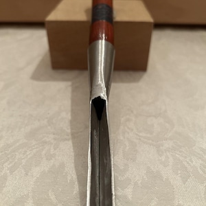 May include: A woodworking chisel featuring a long, sharp metal blade with a concave profile and a cylindrical handle made of reddish-brown wood with a dark band near the center and a metallic cap at the top end of the tool handle.