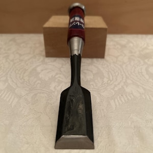 May include: A Japanese wood chisel featuring a wide, flat, dark-colored metal blade and a cylindrical handle made of dark red wood. The handle includes a metal ferrule at the top and a blue label with white Japanese text printed on the side.