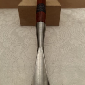 May include: A wood carving gouge featuring a polished metal blade with a curved edge and a cylindrical wooden handle. The handle has a dark brown finish, a metallic cap at the top, and a small black label with white text wrapped around the center.