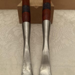 May include: Two metal tools with long, slender, silver-colored shafts and dark brown wooden handles. Each handle features a dark band with the text "Lorentz" printed in a light color. The tools have a flat, slightly curved, wide metal tip.