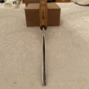 May include: A wood carving gouge with a long, curved metal blade and a light brown wooden handle. The handle features the text SWISS MADE stamped into the wood. The metal shaft is narrow and features a deep, U-shaped channel for carving wood.