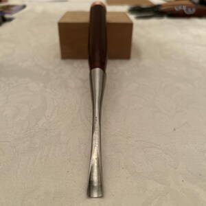 May include: A woodworking chisel with a dark brown wooden handle, a copper-colored ferrule, and a silver-colored blade. The chisel is designed for detailed carving and shaping of wood. The tool is shown against a neutral background.