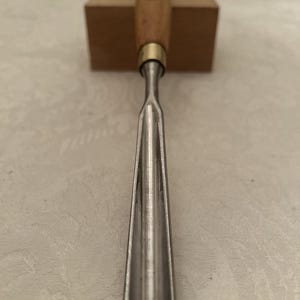 May include: A wood carving chisel with a wooden handle and a metal blade. The chisel is resting on a small wooden block. The handle is light brown, and the blade is silver.