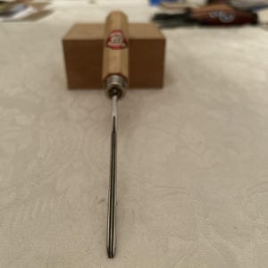 Puede incluir: Una lezna con mango de madera y eje metálico, ideal para trabajos en cuero o manualidades. El mango tiene una etiqueta roja y blanca. La lezna está colocada verticalmente contra un bloque de madera, con la punta hacia abajo.