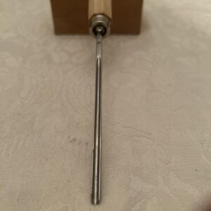 Puede incluir: Una herramienta de cincel de metal y madera con una hoja plateada y un mango de madera. El cincel tiene un eje metálico cilíndrico y una base de madera rectangular. La herramienta está diseñada para trabajar la madera y tallar.