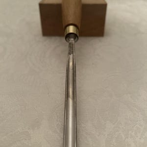 May include: A wood carving chisel with a polished metal blade and a wooden handle. The handle is a light brown color, and the blade is silver. The chisel is resting on a wooden block.