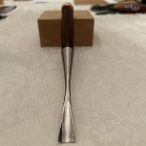 May include: A woodworking chisel with a dark brown wooden handle, a silver-colored metal shaft, and a copper-colored end cap. The chisel is designed for detailed wood carving and shaping.
