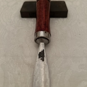 Puede incluir: Un formón para carpintería con un mango de madera rojo oscuro y una virola de metal plateado. La hoja es plateada y muestra signos de uso. Una piedra de afilar gris oscuro y una caja de madera están en el fondo.