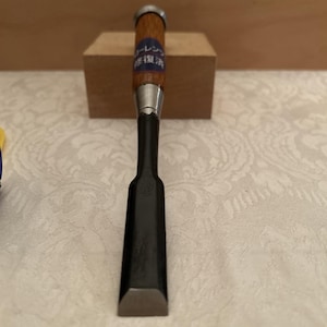 May include: A wood chisel featuring a dark metal blade with a beveled edge and a wooden handle. The handle includes a metal ferrule at the top and a blue circular sticker with white Japanese text that reads "コーレン 修復済".