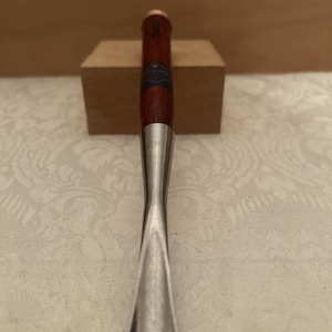 Op de afbeelding: Een houtbeitel met een lang, gebogen metalen blad met een gepolijste afwerking en een donkerbruin houten handvat. Het handvat is voorzien van een klein, donker rond label met tekst bij de bovenkant en een koperkleurige dop aan het uiteinde.