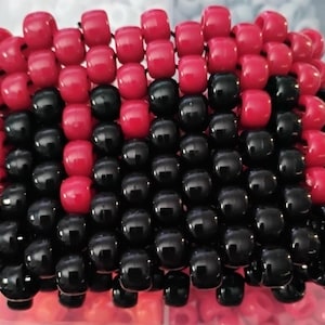 May include: A handmade beaded bracelet featuring a pattern of red and black beads. The bracelet is constructed with small, round beads strung together to form a rectangular shape. The color scheme is a bold contrast of red and black.