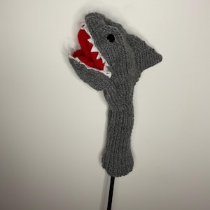 May include: Gray knitted golf club head cover shaped like a shark with a red and white open mouth.