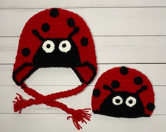 May include: Two crocheted ladybug hats. The larger hat is red with black spots and a black face, white eyes, and red braided ties. The smaller hat is red with black spots and a black face with white eyes. The hats are handmade.