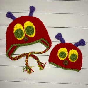 May include: Two crocheted caterpillar hats, one larger than the other, with red bodies, green and yellow eyes, and purple antennas. The hats have green and yellow braided ties.