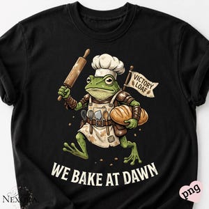 Könnte beinhalten: Schwarzes T-Shirt mit einem Cartoon-Frosch als Bäcker, der einen Nudelholz und ein Brotlaib hält. Der Frosch trägt eine Kochmütze und eine Schürze, darunter der Text "WE BAKE AT DAWN". Eine Flagge zeigt "VICTORY LOAF".