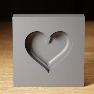 Puede incluir: Un bloque cuadrado gris con una hendidura en forma de corazón. El corazón está tallado en la superficie del bloque.