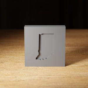 Può includere: Un blocco quadrato grigio con un ritaglio dello stato dell'Indiana. Il blocco è appoggiato su una superficie di legno.