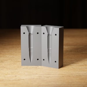 May include: Two-part grey mould for a bullet-shaped object. Each half of the mould has a cavity in the shape of a bullet, with a conical top and a pointed tip. The mould is made of a solid, opaque material.