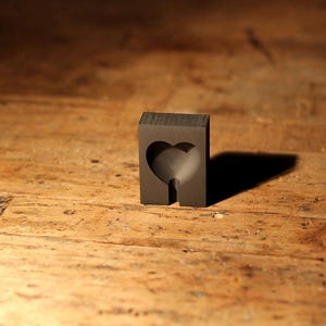 Peut inclure: Un bloc rectangulaire gris foncé avec une découpe en forme de cœur. Le bloc a une extension en forme de tige en bas. L'objet projette une ombre sur la surface en bois.
