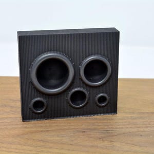 May include: A black rectangular block with five circular cutouts of varying sizes. The block has a textured surface.