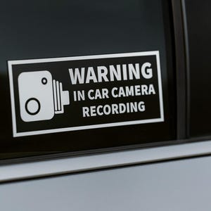 Può includere: Un adesivo rettangolare bianco con bordo nero, applicato al finestrino di un'auto. L'adesivo presenta un'icona di una telecamera e le parole "WARNING IN CAR CAMERA RECORDING" in grassetto bianco.