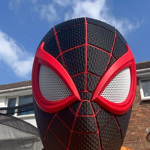 Puede incluir: Una m&aacute;scara de Spider-Man negra y roja con una superficie texturizada y un dise&ntilde;o de telara&ntilde;a roja. Las aberturas de los ojos son blancas con marcos rojos. La m&aacute;scara la lleva una persona con una camiseta naranja.