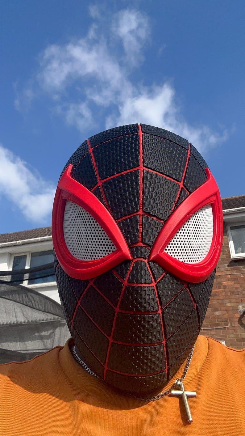 Puede incluir: Una m&aacute;scara de Spider-Man negra y roja con una superficie texturizada y un dise&ntilde;o de telara&ntilde;a roja. Las aberturas de los ojos son blancas con marcos rojos. La m&aacute;scara la lleva una persona con una camiseta naranja.