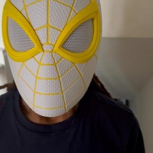 Puede incluir: M&aacute;scara de Spider-Man blanca y amarilla con un dise&ntilde;o de telara&ntilde;a. La m&aacute;scara tiene marcos de ojos amarillos y una cubierta de malla para los ojos. La m&aacute;scara es usada por una persona que lleva una camiseta azul marino.