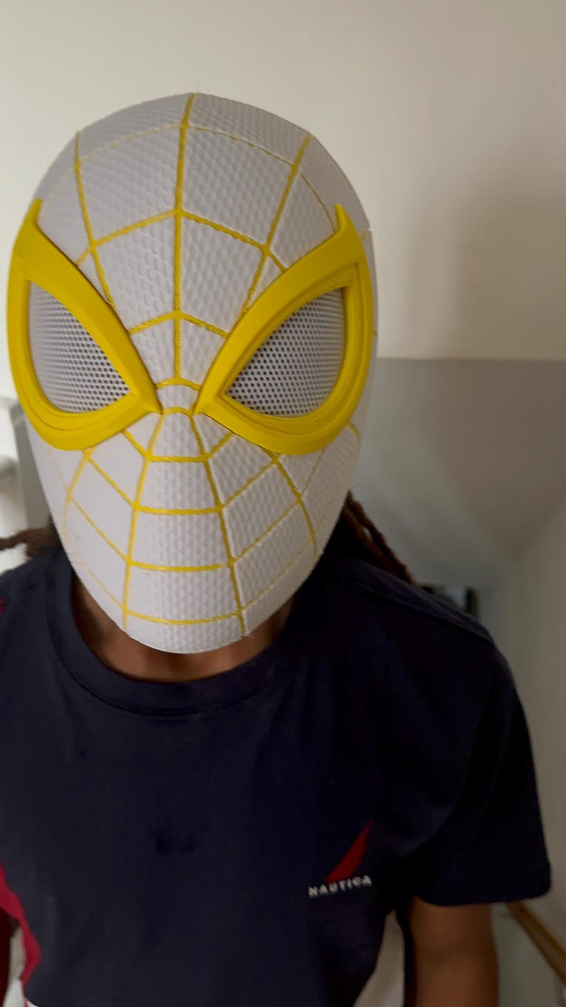 Puede incluir: M&aacute;scara de Spider-Man blanca y amarilla con un dise&ntilde;o de telara&ntilde;a. La m&aacute;scara tiene marcos de ojos amarillos y una cubierta de malla para los ojos. La m&aacute;scara es usada por una persona que lleva una camiseta azul marino.