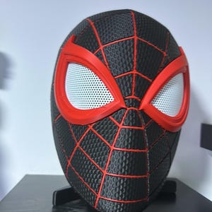 Puede incluir: Una m&aacute;scara de Spider-Man negra y roja con una superficie texturizada y l&iacute;neas rojas de telara&ntilde;a. Las aberturas de los ojos son blancas con un patr&oacute;n de malla, y la m&aacute;scara se muestra sobre un soporte negro. Es un art&iacute;culo de cosplay.