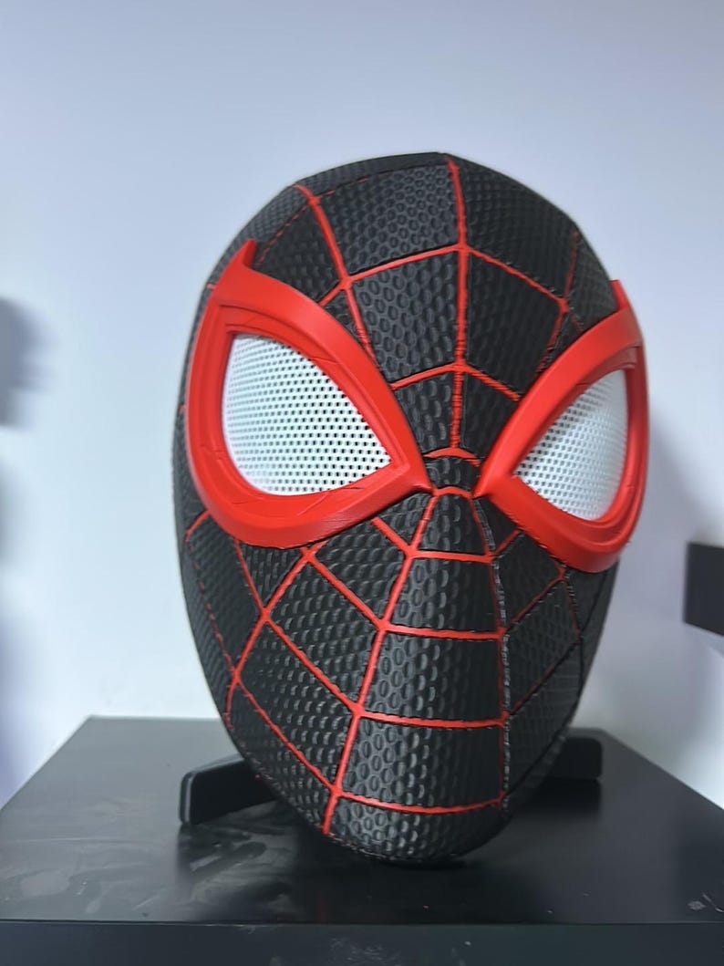 Puede incluir: Una m&aacute;scara de Spider-Man negra y roja con una superficie texturizada y l&iacute;neas rojas de telara&ntilde;a. Las aberturas de los ojos son blancas con un patr&oacute;n de malla, y la m&aacute;scara se muestra sobre un soporte negro. Es un art&iacute;culo de cosplay.