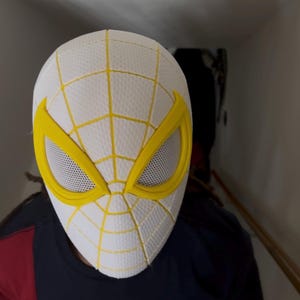Puede incluir: M&aacute;scara de Spider-Man blanca y amarilla con un dise&ntilde;o de telara&ntilde;a. La m&aacute;scara tiene marcos de ojos amarillos y una cubierta ocular de malla. La persona que lleva la m&aacute;scara lleva una camisa azul marino, roja y blanca.