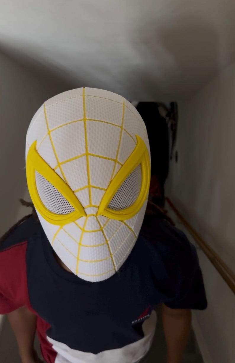 Puede incluir: M&aacute;scara de Spider-Man blanca y amarilla con un dise&ntilde;o de telara&ntilde;a. La m&aacute;scara tiene marcos de ojos amarillos y una cubierta ocular de malla. La persona que lleva la m&aacute;scara lleva una camisa azul marino, roja y blanca.