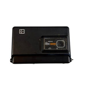 以下が含まれることがあります： 黒いKodak Disc 6000カメラ。カメラは長方形で、左上に正方形のKodakロゴがあります。カメラの前面には、白とオレンジで「KODAK disc 6000」という文字が書かれています。このカメラはヴィンテージ写真アイテムです。